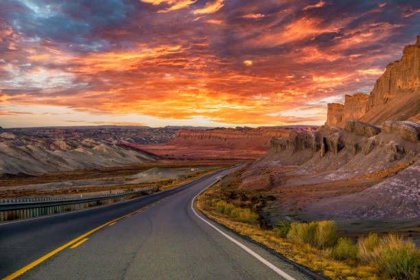 Sunset over Capitol Reef National Park in Utah, where a scenic road winds through dramatic red rock formations and rugged desert terrain. This stunning view captures the beauty of the American Southwest’s vast landscapes and open roads.