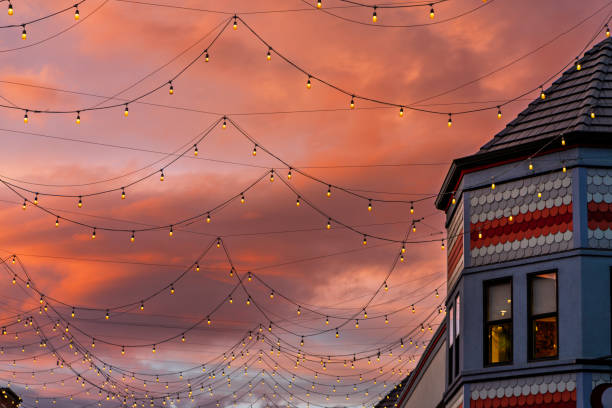 Light Bulbs Strung Across Buildings at Dusk with Sunset Sky - Warm toned light bulbs up against pink and orange sunset sky with clouds lit with warm light.