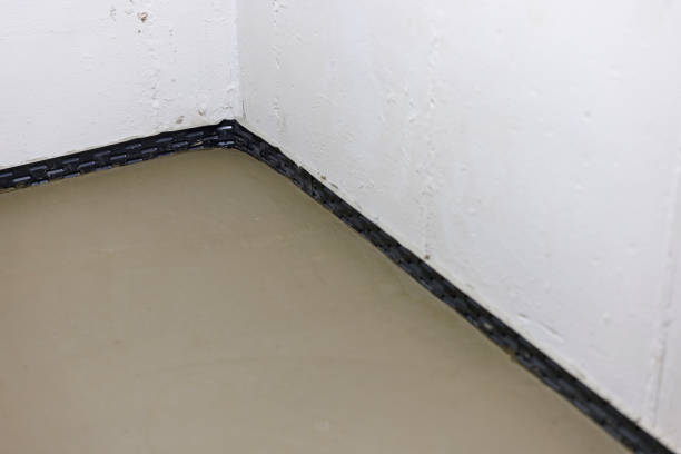 Level concrete floor installation meets black waterproof membrane at the wall junction of a basement corner. The protective barrier creates a water-tight seal between floor and wall, part of a comprehensive moisture control system for below-grade spaces. Fresh concrete work demonstrates professional-grade foundation waterproofing techniques