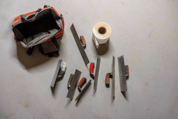 Professional-grade plastering equipment and finishing tools arranged on concrete surface, including specialty trowels, putty knives, tool bag, and construction materials prepared for basement waterproofing and drainage system installation