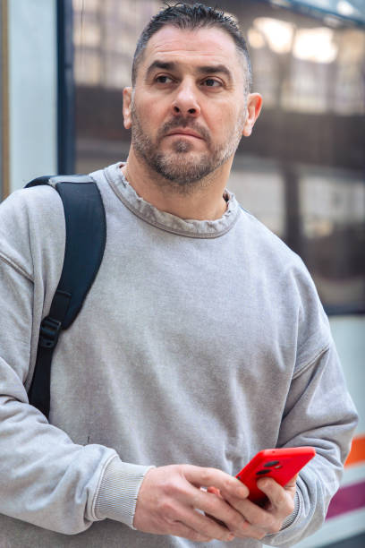 Serious man wearing a gray sweatshirt holding a red smartphone while looking around at a train station. His expression conveys anticipation, travel, and modern connectivity.