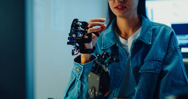 istock Closeup Young female engineer working late gestures towards robotic arm in tech lab at night. Machine learning, computer manufacturing, innovation and creativity. AI and Robotics Development. 2197913315