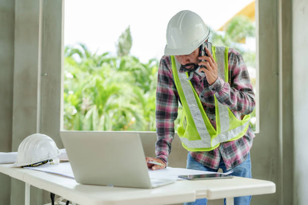 istock Architect wearing safety helmet and reflective vest talking on mobile phone while reviewing blueprint on construction site. Laptop and safety equipment placed on table near open window. 2197917199