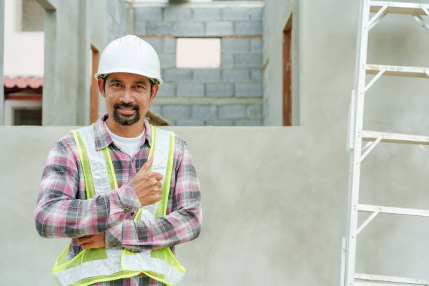 istock Construction engineer wearing safety helmet and reflective vest smiling while standing at residential site. Unfinished house with concrete framework and building materials visible in background. 2197917229