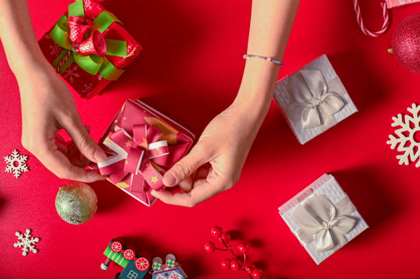 istock Two hands hold tiny red gift boxes on a festive red background. Around them are larger wrapped gifts, snowflakes, a toy train, ornaments, and berries, creating a bright and cheerful Christmas scene. 2197917358