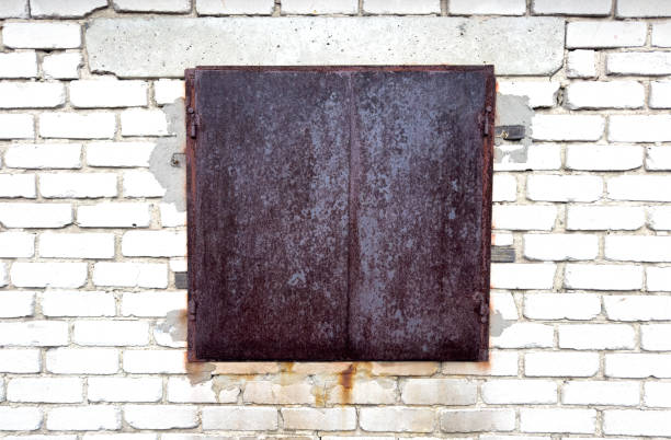 A window with a metal door is on a brick wall. The door is rusted and the window is empty