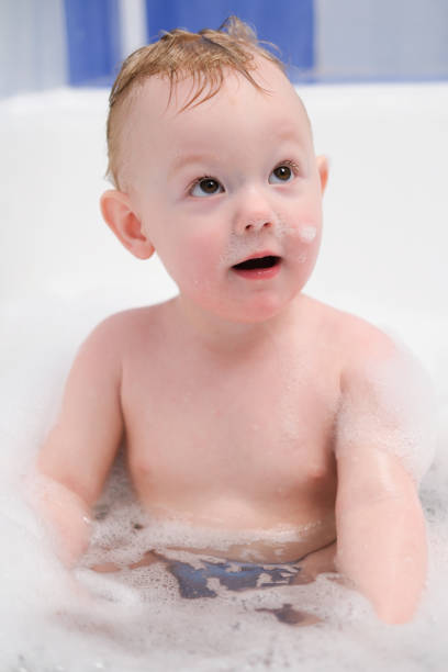 A cute baby is sitting in a white bathtub, smiling and holding a yellow toy, enjoying playful bath time under bright natural light. Captures innocence, joy, and happy moments of childhood.