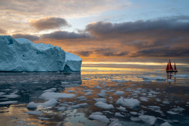 red sailboat sailing among floating icebergs in Disko Bay glacier with orange sunset during polar summer midnight solar season. Ilulissat, Greenland