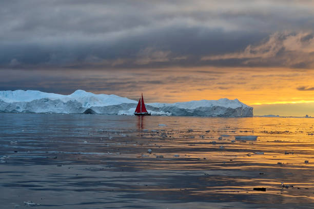 red sailboat sailing among floating icebergs in Disko Bay glacier with orange sunset during polar summer midnight solar season. Ilulissat, Greenland