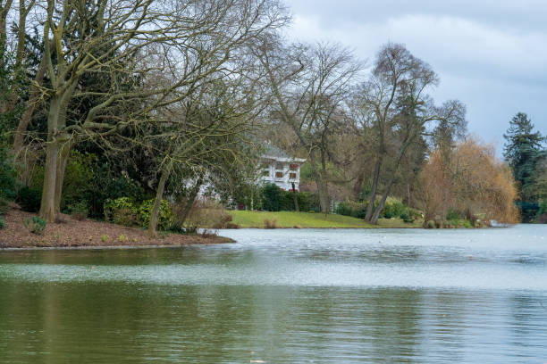 A tranquil setting of a lake surrounded by bare trees, reflecting a quaint house in the distance, evoking peace on a cool, overcast afternoon.