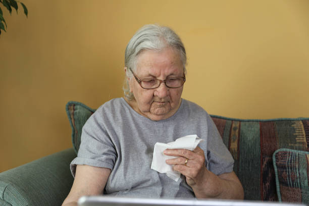 The image shows an elderly woman with gray hair and glasses sitting on a couch in the living room. She is dressed in a loose gray dress and holds a tissue in her hand while looking at an open laptop on her lap. Her face reflects emotions, possibly sadness or concern.