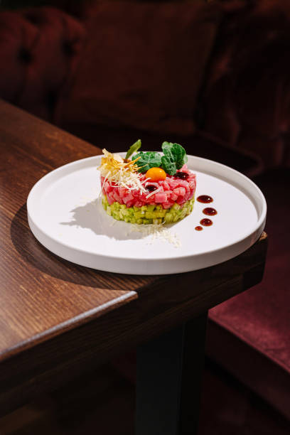 A beautifully crafted dish showcases layers of fresh vegetables and proteins, adorned with herbs and sauces. The plate is elegantly set on a wooden table, creating a warm dining atmosphere.