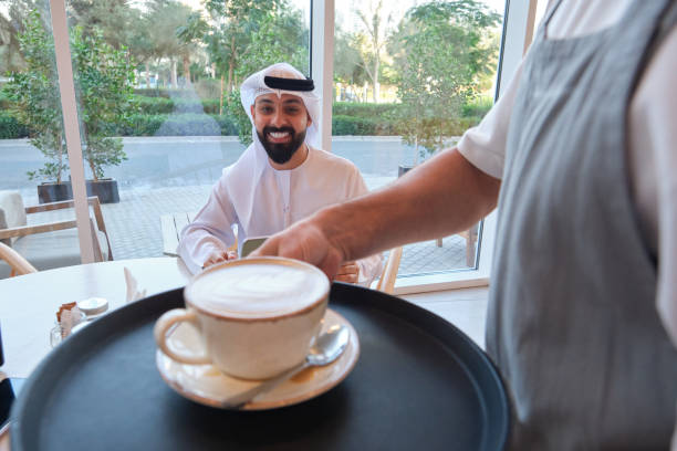 Smiling Saudi man receiving coffee while sitting at cafe. Waiter is serving cappuccino to customer. He is wearing traditional clothes.