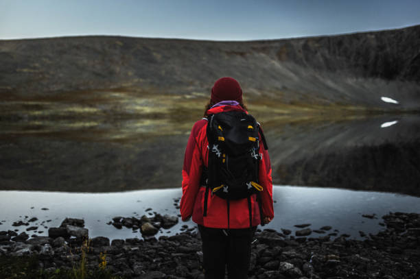 A person stands by a calm and reflective mountain lake, wearing hiking gear, surrounded by a rugged wilderness atmosphere, perfect for outdoor adventures and nature exploration.