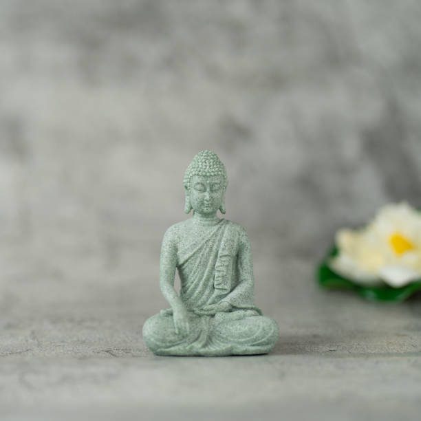 istock A small Buddha statue is sitting on a gray surface. The statue is made of stone and has a serene expression on its face. in the background an out-of-focus lotus flower is visible. 2197936185