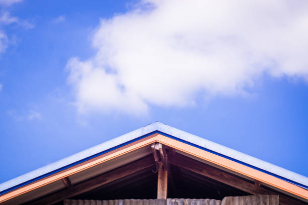 The image captures the gable of a house roof with a clear blue sky and fluffy white clouds in the background. The roof has a distinct triangular shape with blue trim and visible wooden support beams, creating a striking contrast between the architectural structure and the natural sky.