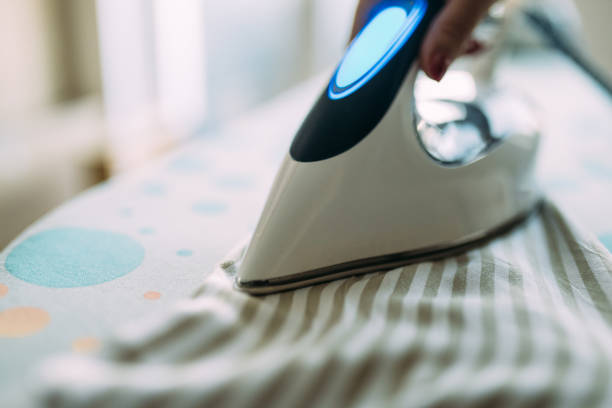 A hand holding a steam iron over an ironing board in a sunlit, cozy room, showcasing domestic chores and daily routine, with the steam creating a dynamic touch to the composition