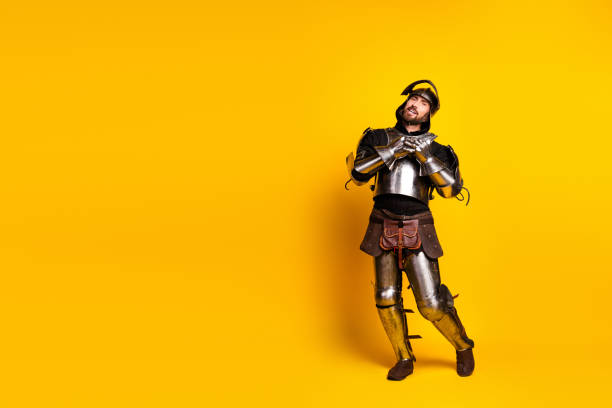 A man dressed as a medieval knight in shining armor strikes a whimsical pose, set against a solid bold yellow background exuding charm and historical essence.