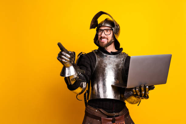 istock Young man in medieval armor pointing and holding a laptop with a cheerful expression on a vibrant yellow background. 2197943403