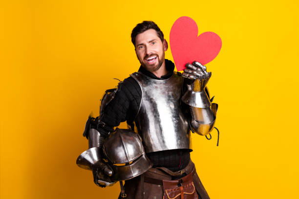 istock Man in medieval knight armor holding a heart prop on bright yellow background 2197943423