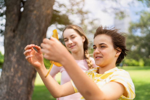 Two children, one in blue and one in red, playfully set up a rope between trees in a wooded area.  They seem to be enjoying a sunny day outdoors.
