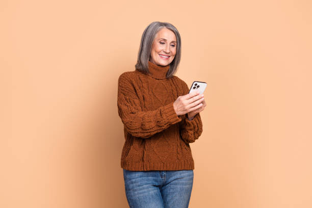 istock Mature woman in a cozy brown sweater smiling while using a smartphone against a neutral beige background 2197944507
