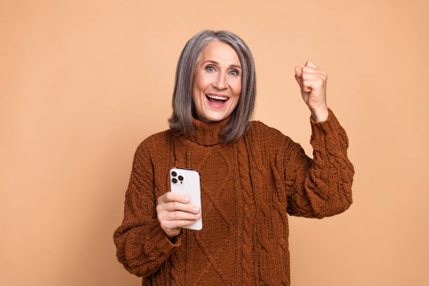 istock Smiling grey-haired woman celebrating success while holding a smartphone, wearing a brown sweater with beige background. 2197944524