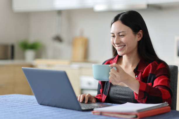 Happy asian woman drinking and e-learning using laptop Happy asian woman drinking and e-learning using laptop in the kitchen at home high school enrollment stock pictures, royalty-free photos & images