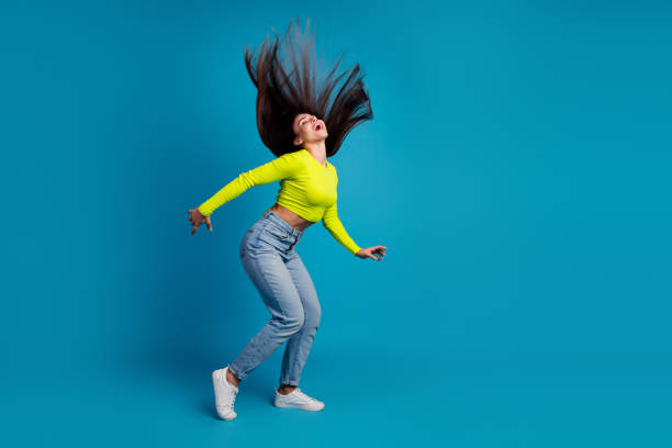 A trendy young woman in casual fashion dances gracefully, flipping her long brunette hair elegantly. The bright yellow top contrasts beautifully with the blue backdrop, emphasizing her joyful and confident demeanor.