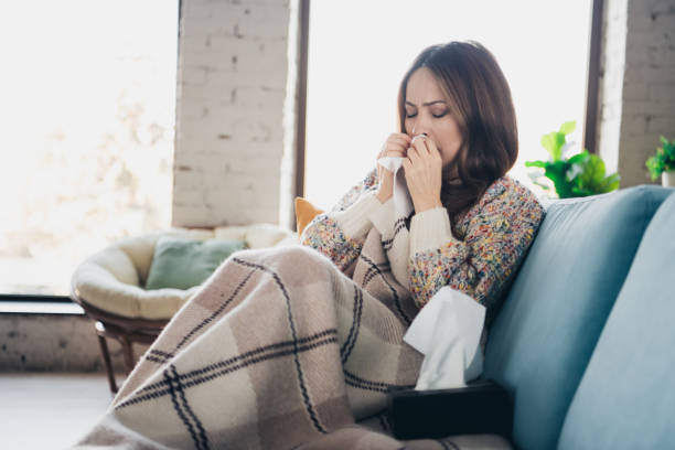 istock Young woman sitting on couch indoors sneezing into tissue under warm blanket 2197952542