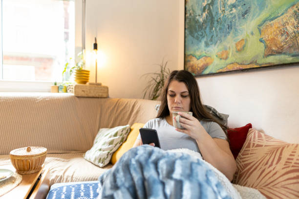 istock Woman in her living room drinking, lying sick and using a mobile device. Wrapped in a cozy blanket, she was placed on the couch. Copy space 2197952825