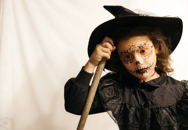 A young girl dressed as a witch with skull face paint, holding a broom and looking serious in an indoor setting.
