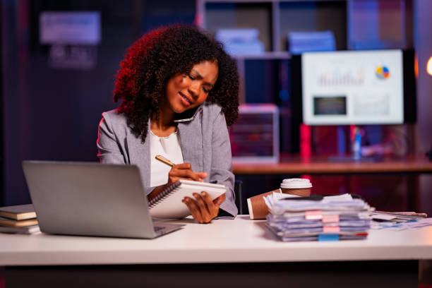 istock African female employee working night in office with bokeh lights to work late on project deadline using smartphone technology and laptop, graph document 2197955918