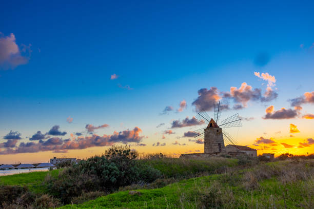 A picturesque windmill stands against a vibrant sunset in Southern Italy. The sky is painted with colorful clouds, while lush greenery surrounds the historic structure, capturing the serene beauty of this enchanting landscape.