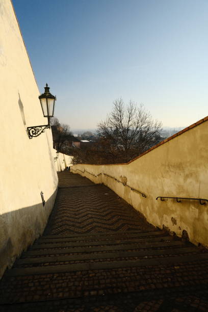 A winding staircase descends along a weathered white wall, lined with a vintage lantern, offering views of surrounding trees and distant landscape beneath a bright sky.