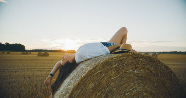 A young Caucasian woman joyfully reclines on a sunlit haystack in a vast, golden field. Dressed casually, she smiles warmly, embodying freedom and peace against the serene countryside sunset.