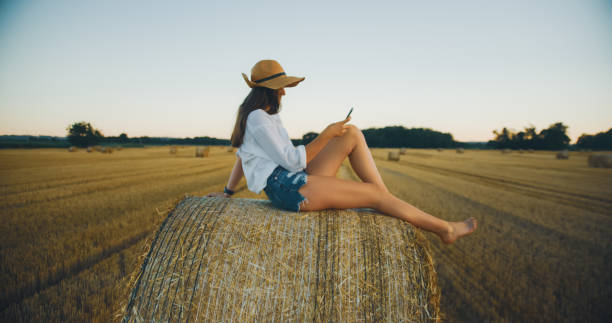 A carefree Caucasian young adult female sits on a haystack, using her smartphone. She wears a casual outfit with a hat, in a serene countryside field during sunset.