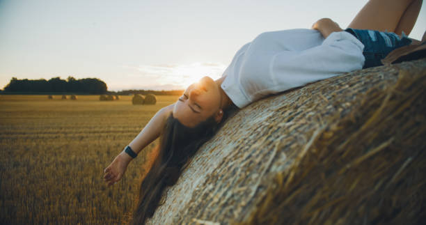 A carefree Caucasian young adult female in casual summer attire relaxes joyfully on a large haystack in a serene, sunlit countryside field during sunset.