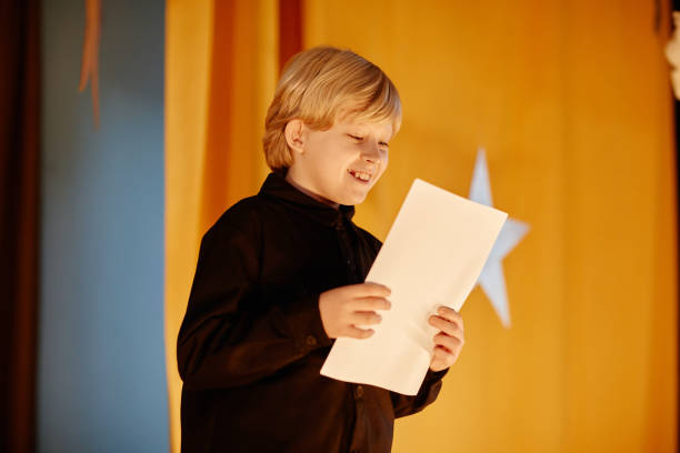 Smiling young boy holding paper while standing on stage during school performance, with a large star visible in background