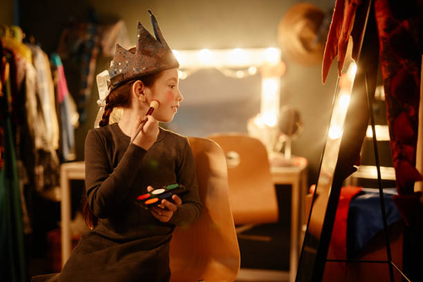 Young girl wearing costume applying makeup in cozy dressing room surrounded by garments and bright light. Engaging in creative play and imagination