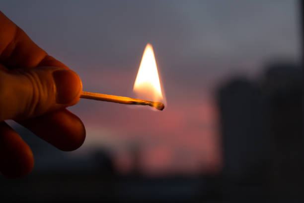 Close-up of a hand holding a lit match. The hand, which is a rich, warm brown tone, is clearly visible, with the fingers and thumb encircling the matchstick.  The matchstick is a light brownish-tan color, and a bright flame is burning at its tip. The flame is a clear orange-yellow. The background is out of focus, showcasing a dusky, purplish-pink sky merging into a dark, almost black background. Hints of buildings or structures are visible in the far background. The depth of field focuses on the hand and match, blurring the background, creating a sense of depth.
