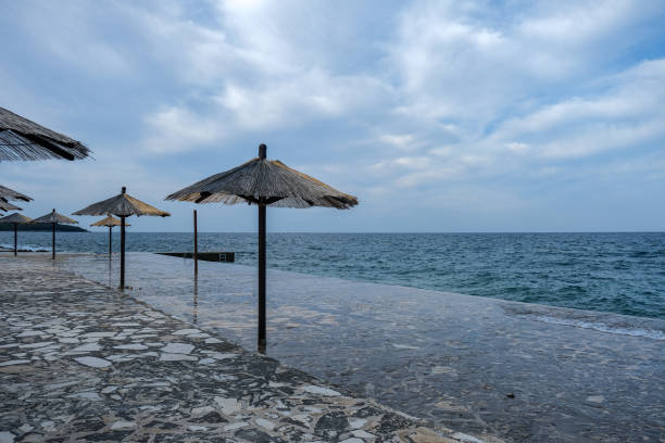 A row of thatched umbrellas stands along a coastal walkway, overlooking the sea under a cloudy sky. The stone pavement appears wet as waves reach the shore, creating a serene seaside scene.