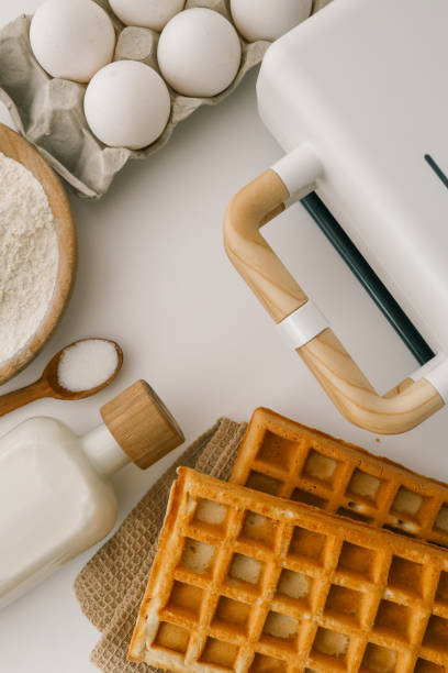 istock Ingredients for homemade waffles arranged on a white surface, including flour, sugar, eggs, and milk. A waffle maker with wooden handles is visible in the frame 2198029063