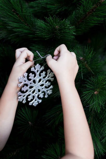 Child hanging silver snowflake ornament on green Christmas tree branches.