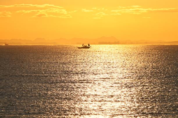 A scene of dawn on a Japanese beach. The silhouettes of fishing boats, birds, surfers and people walking are bathed in the colors of the rising sun, making it a very beautiful sight.