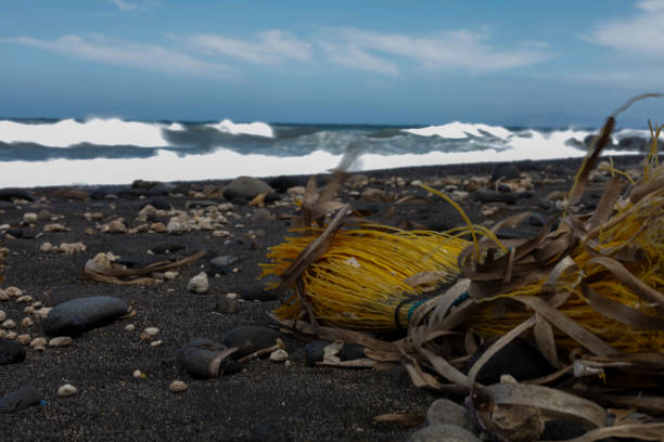 A coastal scene with scattered debris, featuring bright yellow garbage broom remnants contrasting against the dark volcanic sand. Waves crash in the background under a partly cloudy sky, creating a dramatic and reflective mood.