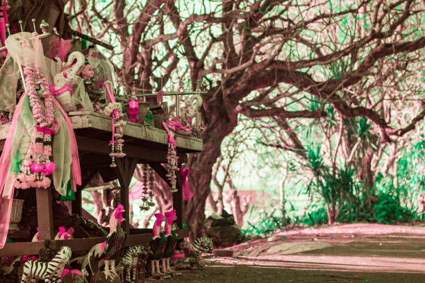 The image features a scene with a table adorned with various decorative items, including floral garlands and perhaps offerings, set against a backdrop of thick, twisted trees. The color palette has a surreal quality, with shades of green and pink. In front of the table, there are decorative elements that resemble animals, likely roosters.