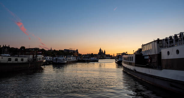 A serene sunset view captured along the IJ River in Amsterdam. Boats are moored on both sides of the river, framing the glowing horizon in the background. The soft colors of the sky, transitioning from deep blue to warm orange, complement the reflective water. The silhouette of the Basilica of Saint Nicholas can be seen in the distance, adding a historical element to the peaceful waterscape.