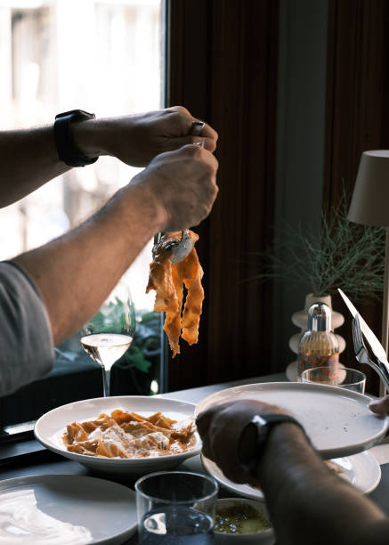 Diners enjoying pappardelle pasta in an Italian restaurant, hands lifting fresh pasta from a creamy tomato-based dish, elegant dining experience with warm ambiance and stylish table setting