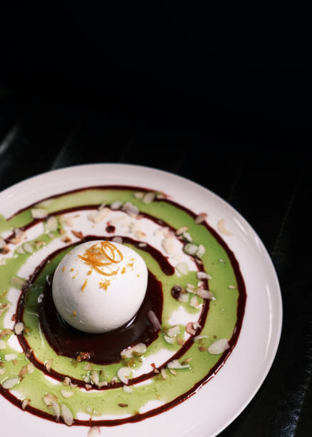 A beautifully plated burrata cheese dish featuring a creamy, delicate burrata ball at the center, surrounded by swirls of vibrant green basil pesto and rich balsamic reduction. The dish is garnished with crushed nuts and delicate herbs, adding texture and visual appeal. Presented on a sleek white plate, the composition stands out against a dark background, evoking a sophisticated Italian dining experience. Ideal for showcasing fine dining, gourmet cheese dishes, and modern Italian cuisine.